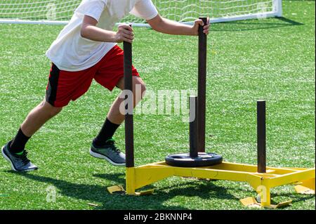 Side view of a high school boy pushing a yellow sled with weight on it for resistance training Stock Photo