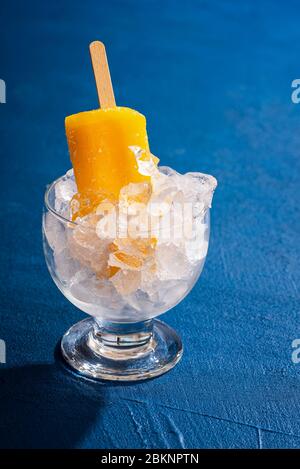 Refreshing orange popsicle cooling in a glass with ice on a classic blue table.  Cold tropical dessert. Summer sweets. Orange ice cream on a stick. Stock Photo