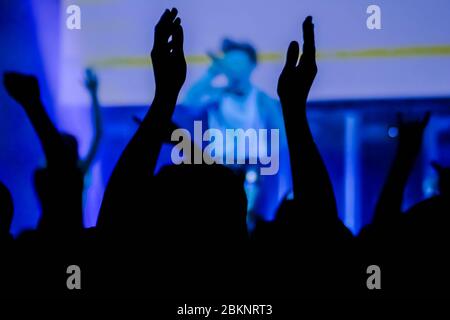 Man silhouette partying, cheering, raising hands up and clapping at rock concert in front of stage of nightclub. Bright colorful blue stage lighting Stock Photo