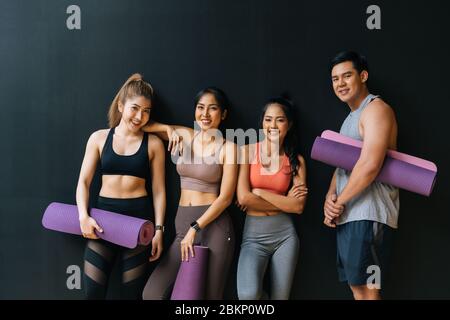 Happy smiling man and women looking at camera altogether in gym. Group of young people relaxing in gym after workout training with black background. Stock Photo