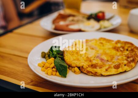 Omelet with ham, peas and corn in a cafe. Delicious omelet breakfast with cheese and peas. Omelet with vegetables on a white plate in a cozy cafe Stock Photo