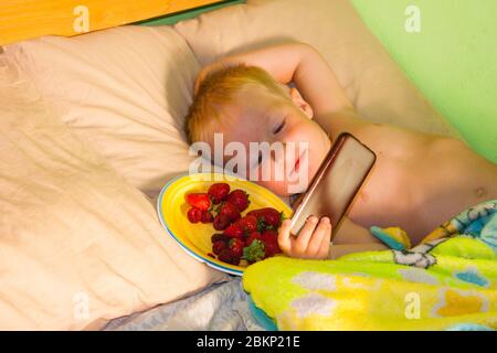 Morning at the cottage - a little boy with blond hair and blue eyes lies on the bed, happily squinting at the sunlight and watching cartoons in the phone. Next to it is a plate with summer berries - strawberries and raspberries. Summer vacation childhood happiness Stock Photo