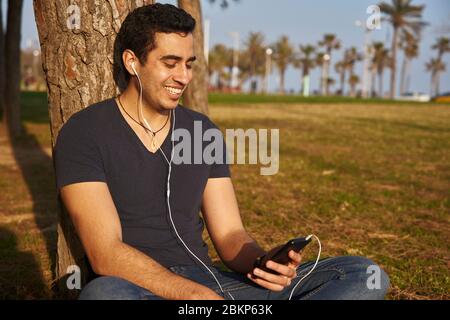 A man sitting against a tree trunk listening to something through earphones and a mobile phone. Stock Photo