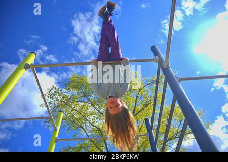 Beautiful blond girl with long hair playing in playground. young girl hanging upside down in a park. Stock Photo
