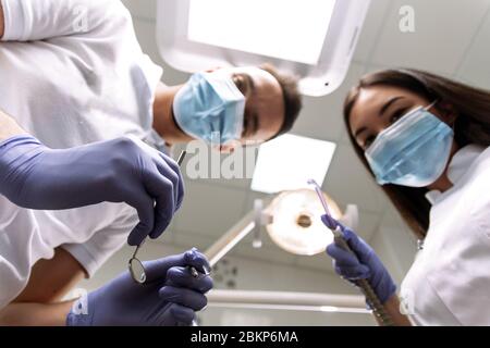 The dentist and assistant bent over the patient to examine the teeth, treat them or create a denture. A man and a woman are holding examination instruments, a saliva tube and looking at the patient. Stock Photo