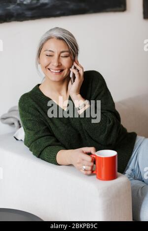 Image of beautiful adult asian woman talking on cellphone and drinking coffee while sitting on sofa at home Stock Photo