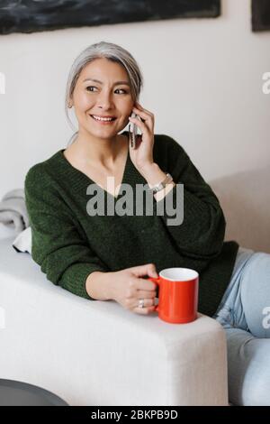 Image of beautiful adult asian woman talking on cellphone and drinking coffee while sitting on sofa at home Stock Photo