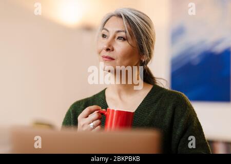Image of positive pleased mature beautiful grey-haired woman using laptop computer indoors drinking coffee. Stock Photo
