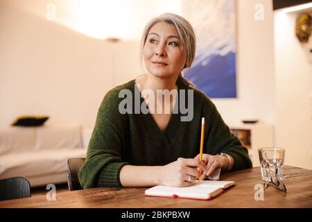 Image of gorgeous concentrated mature beautiful grey-haired woman indoors writing notes. Stock Photo