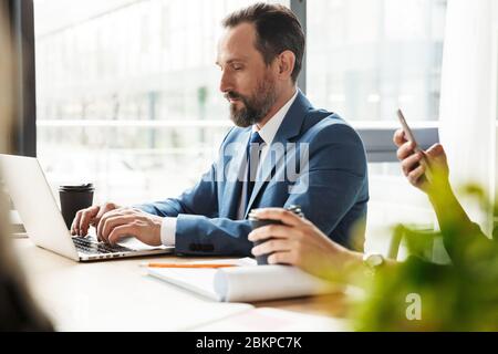 Photo of adult focused colleagues working with laptop and smartphone while sitting at table in office Stock Photo