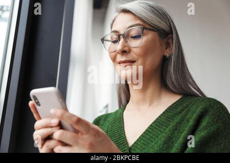 Photo of gray-haired focused businesswoman in eyeglasses using cellphone while working in office Stock Photo