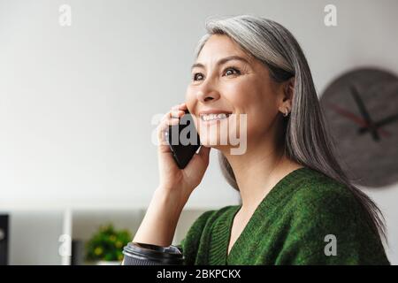 Photo of gray-haired smiling businesswoman talking on cellphone while working in office Stock Photo