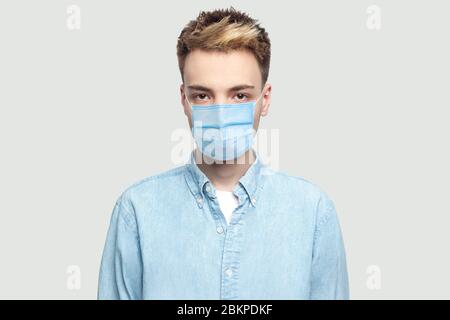 Portrait of calm serious handsome young man with surgical medical mask in light blue shirt standing and looking at camera with serious face. indoor st Stock Photo