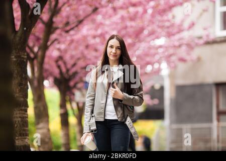 Girl in stylish outfit smiling and holding coffee on sakura background. Portrait of woman in jacket with cup of tea. Stock Photo