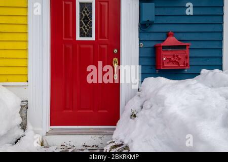 Red door of a building with blue and yellow wooden clapboard walls. The house has a red metal mailbox. There are drifts of white snow in front.