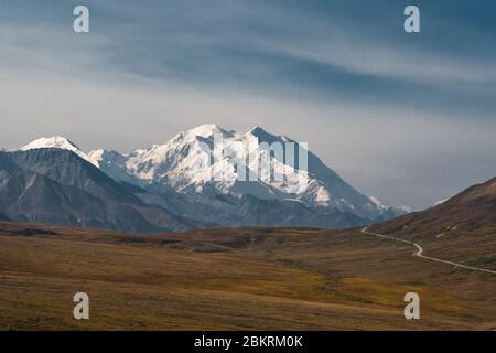 A gravel road through the autumn colored tundra vanishing into the mountains with Mount Denali-McKinley in the background