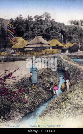 [ 1900s Japan - Japanese Boys Playing in the Countryside ] —   Three country boys playing in a small stream. In the background are houses with thatched roofs.  One of the boys has a pole with Koinobori (carp streamers) to celebrate Tango no Sekku, popularly known as “Boys’ Day”. It was celebrated on the 5th day of the 5th moon in the lunar calendar.  20th century vintage postcard. Stock Photo