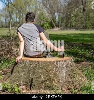 bottle of alcohol in the hand of a man sitting on a stump in the woods Stock Photo