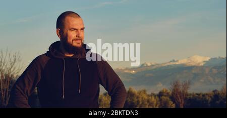 Man in casual hoodie pose against Sierra Nevada mountain ridge background. Traveller male standing outdoors during sundown looking in distance Stock Photo