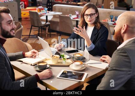 Group of business colleagues sitting at table with coffee cups in cafe and discussing business ideas using portable devices Stock Photo