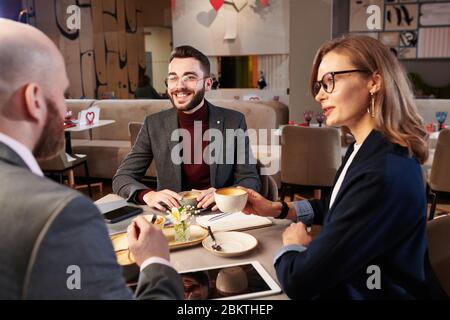 Young business people sitting at table with gadgets and discussing strategy of project implementation while drinking coffee in restaurant Stock Photo