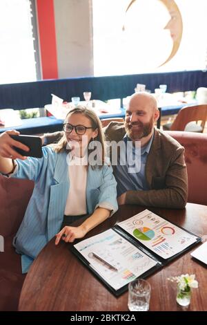 Positive colleagues in jackets sitting at table with financial documents in cafe and taking selfie on smartphone Stock Photo