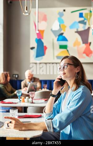 Content confident lady entrepreneur in eyeglasses sitting at table in art cafe and talking on mobile phone Stock Photo