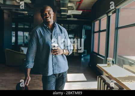 Relaxed businessman standing in office holding a coffee. African male executive having a coffee break at work. Stock Photo