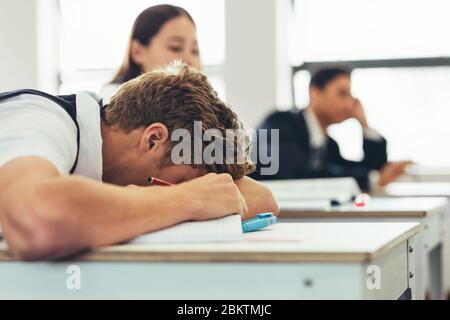 Bored male high school student sleeping during the lecture in the classroom. Schoolboy sleeping on desk in classroom with classmates in background. Stock Photo