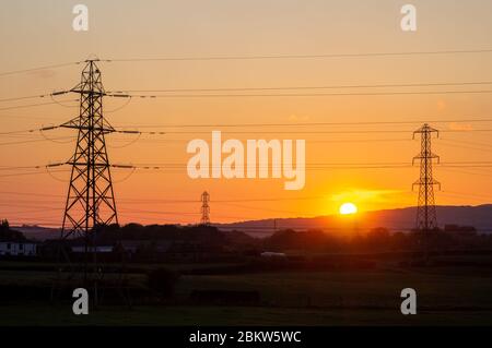 Sun setting between pylons in field - energy power distribution transmission electric electricity Stock Photo