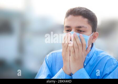 Portrait shot of a coughing man struggling wearing a medical protective mask posing wearing a blue colored corporate shirt over blurred background. Stock Photo
