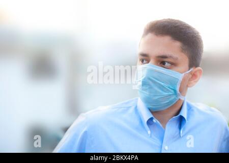 Portrait shot of a young man in blue colored shirt and wearing a surgical mask or a procedure mask with blurred background. Stock Photo