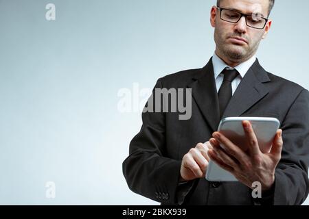 Serious young businessman in black suit and glasses, holding tablet computer and looking at the screen. Low angle front portrait against clean backgro Stock Photo
