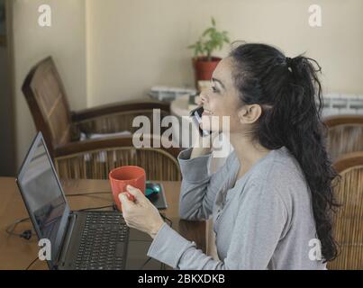 Brunette woman teleworking at home for COVID-19, looking at the computer talking on the mobile Stock Photo