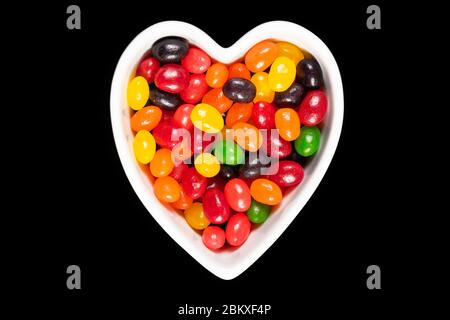 Assorted colorful jelly beans, sweet candy background with a heart-shaped bowl isolated on black. Kids' junk food. Stock Photo