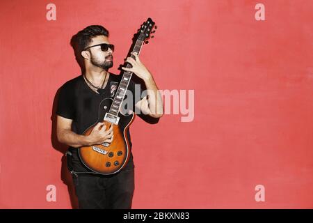 Closeup of one handsome passionate expressive cool young brunette rock musician men playing electric guitar standing against red background Stock Photo