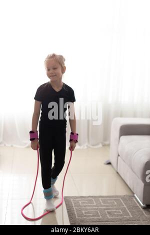 Happy little girl jumping over the rope at home during the isolation. The bright sun illuminates the room. Stock Photo