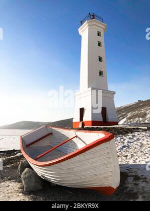 Picture of old lighthouse and wooden boat on the seashore of Magadan, Russia. Tall white building and boat with red stripes. Clear blue sky and ice on the sea in winter. Stock Photo