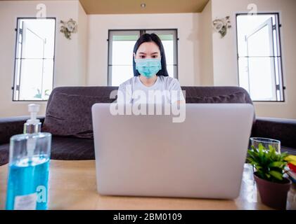 Asian woman cute beautiful is wearing protective mask and using a notebook computer and sitting on sofa for working from home. She is quarantined at h Stock Photo
