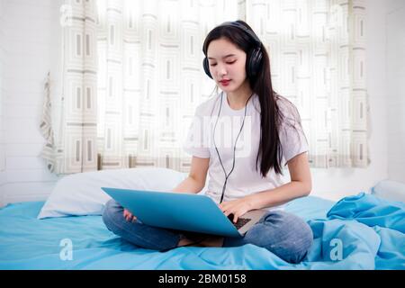 Asian happy young woman wear headphones and listening music song and watching website for shopping online from laptop  on bed at home Stock Photo