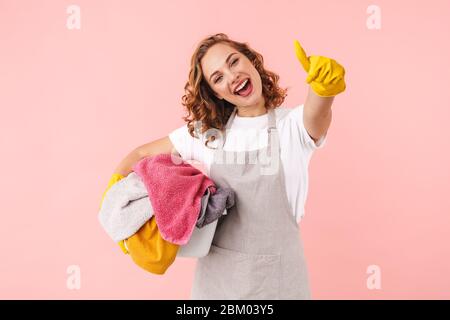 Picture of a positive happy young woman housewife in gloves isolated over pink wall background holding laundry showing thumbs up. Stock Photo