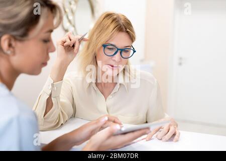 Cosmetology specialist answering clients questions about beauty procedure, mature woman in glasses attentively reading agreement Stock Photo