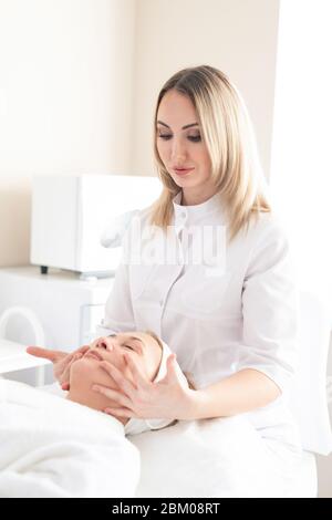 Blonde cosmetologist in white coat sculpting face of mature woman with massage at beauty treatment procedure Stock Photo