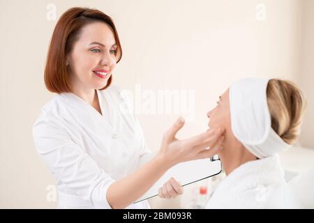 Smiling redhead cosmetologist with clipboard checking face skin of mature client before cosmetic procedure Stock Photo