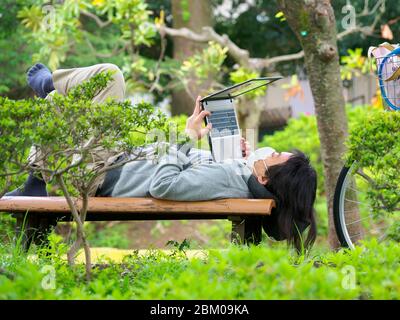 Man using a laptop computer while lying on a bench in a park in Tokyo, Japan. Stock Photo