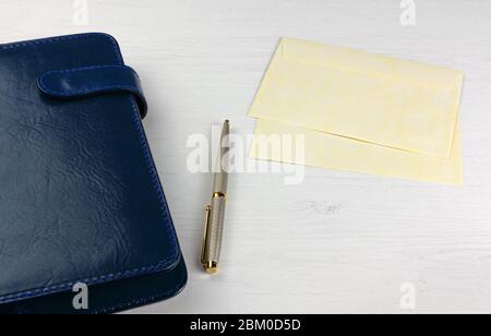 An elegant pen, a daily planner with a blue leather cover and two yellow envelopes on a white wooden desk Stock Photo