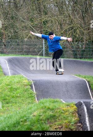 Young man in sportswear riding on skateboard with arms outstretched along hilly asphalt road performing tricks in skate park in daylight Stock Photo