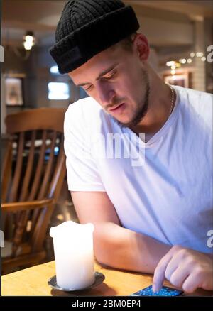 Trendy thoughtful crop male in casual clothes and hat focusing on screen and interacting with smartphone while sitting at table in contemporary bar Stock Photo