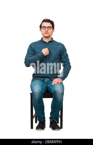 Full length portrait of perplexed businessman, seated on a chair  points index finger to himself, looking confused to camera isolated on white backgro