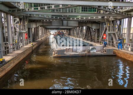 A barge loaded with coiled wire rod lies in the flooded trough of the Niederfinow boat lift, which has bridged a height difference of around 36 metres between the Oder-Havel Canal and the Oder since it opened in 1934. Ring buoys hang from the walkways on either side, and a man in blue work clothes stands at the right edge of the trough Stock Photo